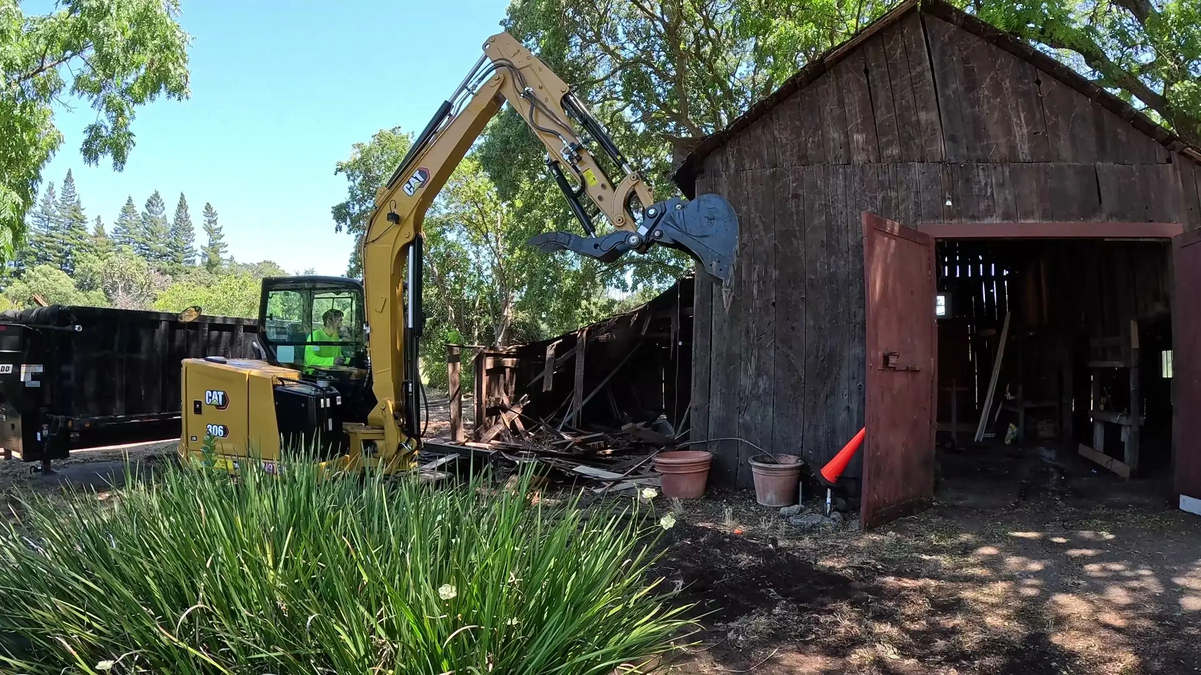 Yellow CAT excavator with raised arm beside a weathered wooden barn and a black dump trailer