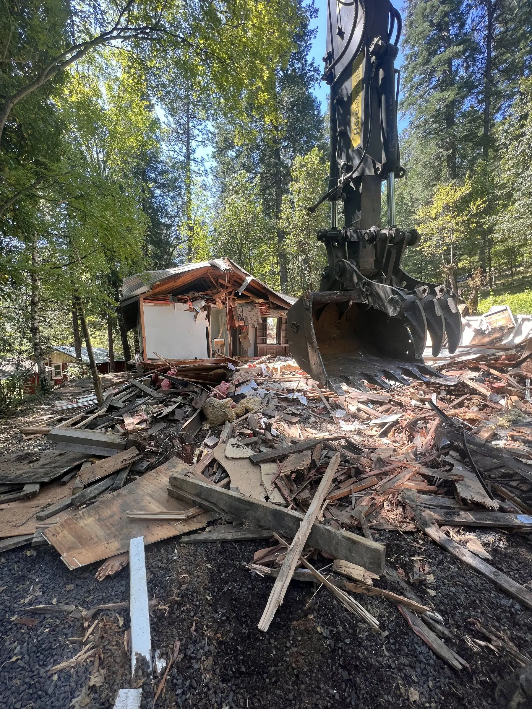Excavator grapple over a pile of house debris among tall pines